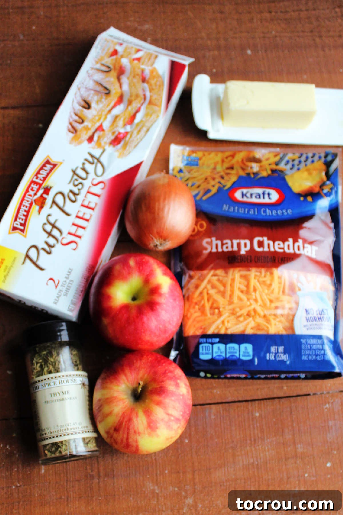 Essential ingredients for Apple Cheddar Pinwheels laid out: a sheet of puff pastry, fresh apples, diced onion, shredded cheddar cheese, a stick of butter, and a sprig of fresh thyme.