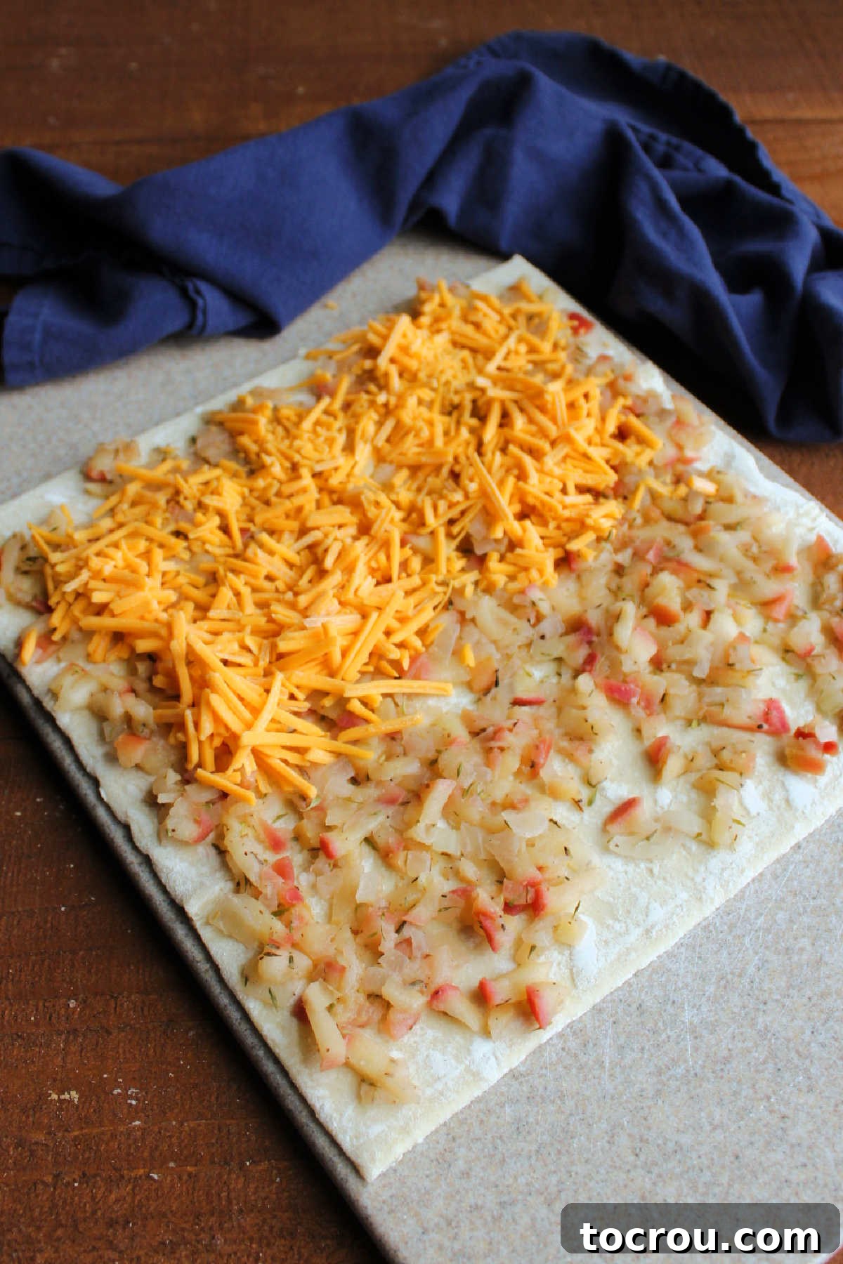 A sheet of light brown puff pastry dough spread flat, generously topped with the cooled apple mixture and a blanket of shredded sharp cheddar cheese, ready for rolling.
