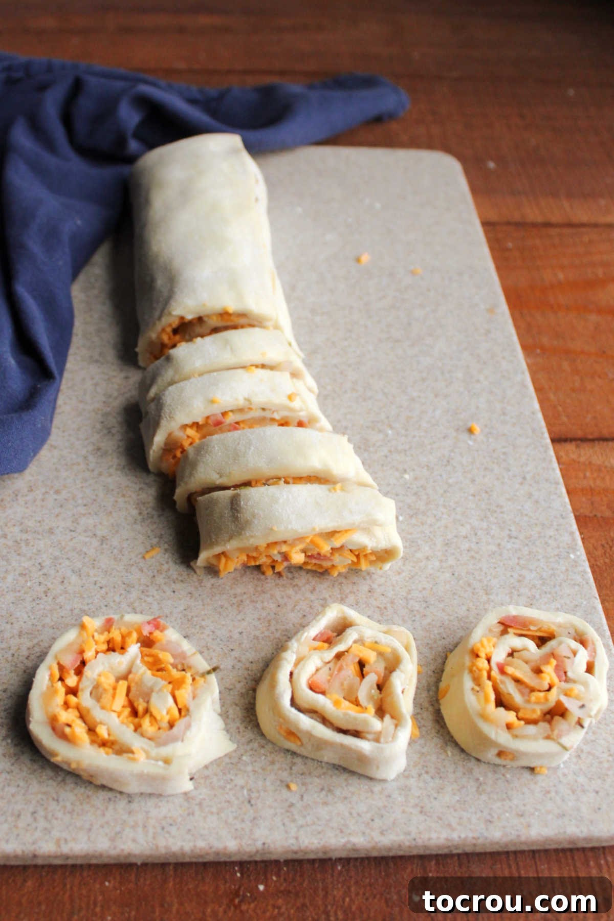A rolled log of puff pastry, filled with apple and cheddar, being carefully sliced into uniform half-inch pinwheel pieces on a cutting board.