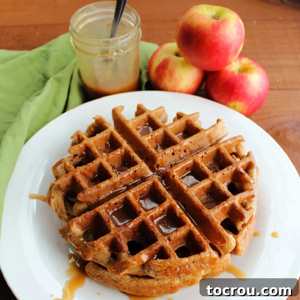 Golden-brown apple cider Belgian waffles drizzled generously with homemade caramel sauce, resting beside a rustic pile of fresh, ripe apples and a light dusting of cinnamon.