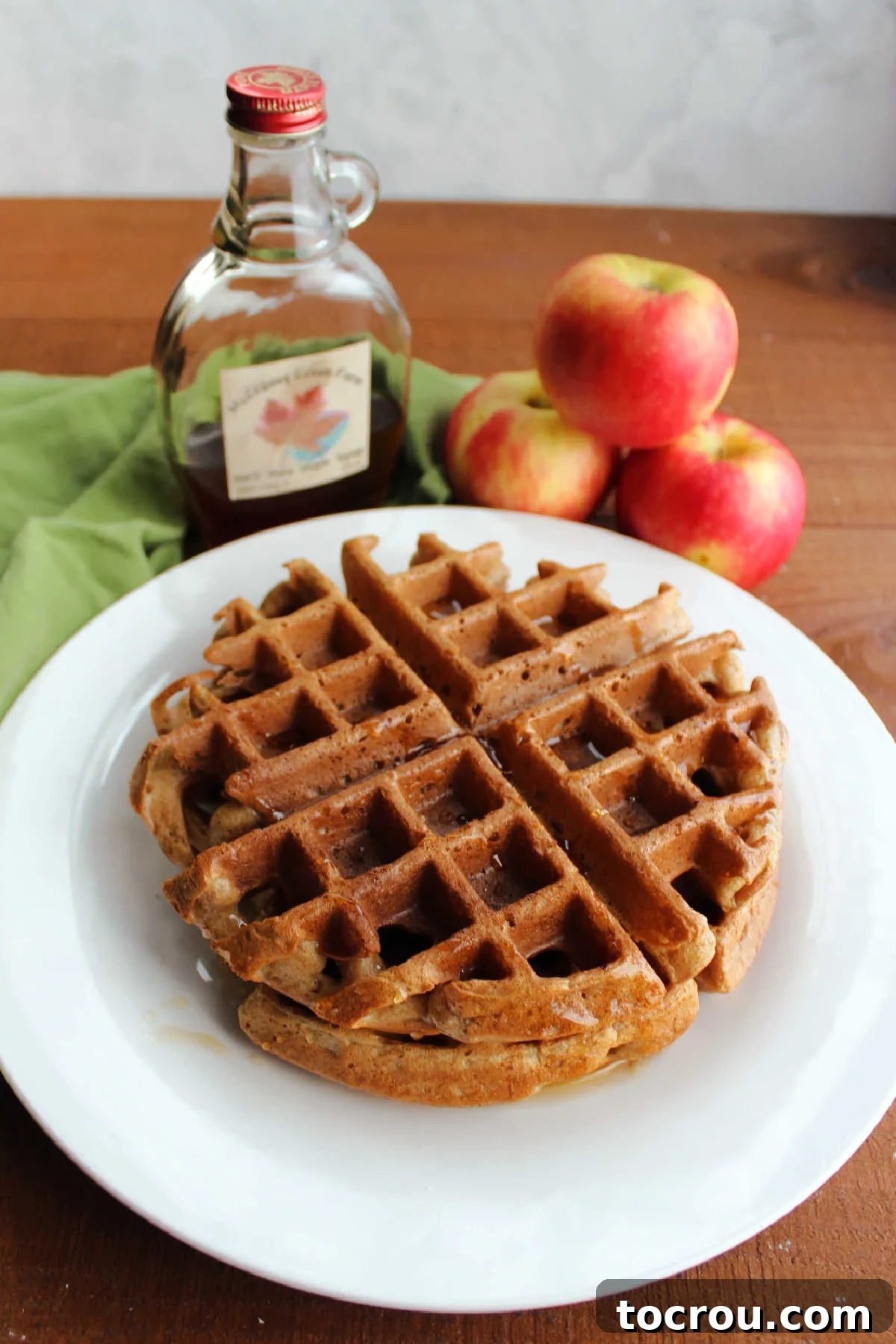 A stack of golden apple cider waffles next to a bottle of maple syrup and fresh apples, evoking a classic fall breakfast scene.