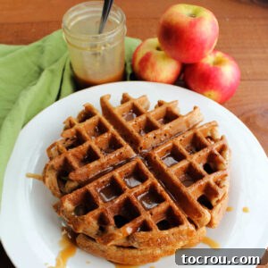 A stack of golden apple cider Belgian waffles drizzled with caramel sauce, next to fresh apples.