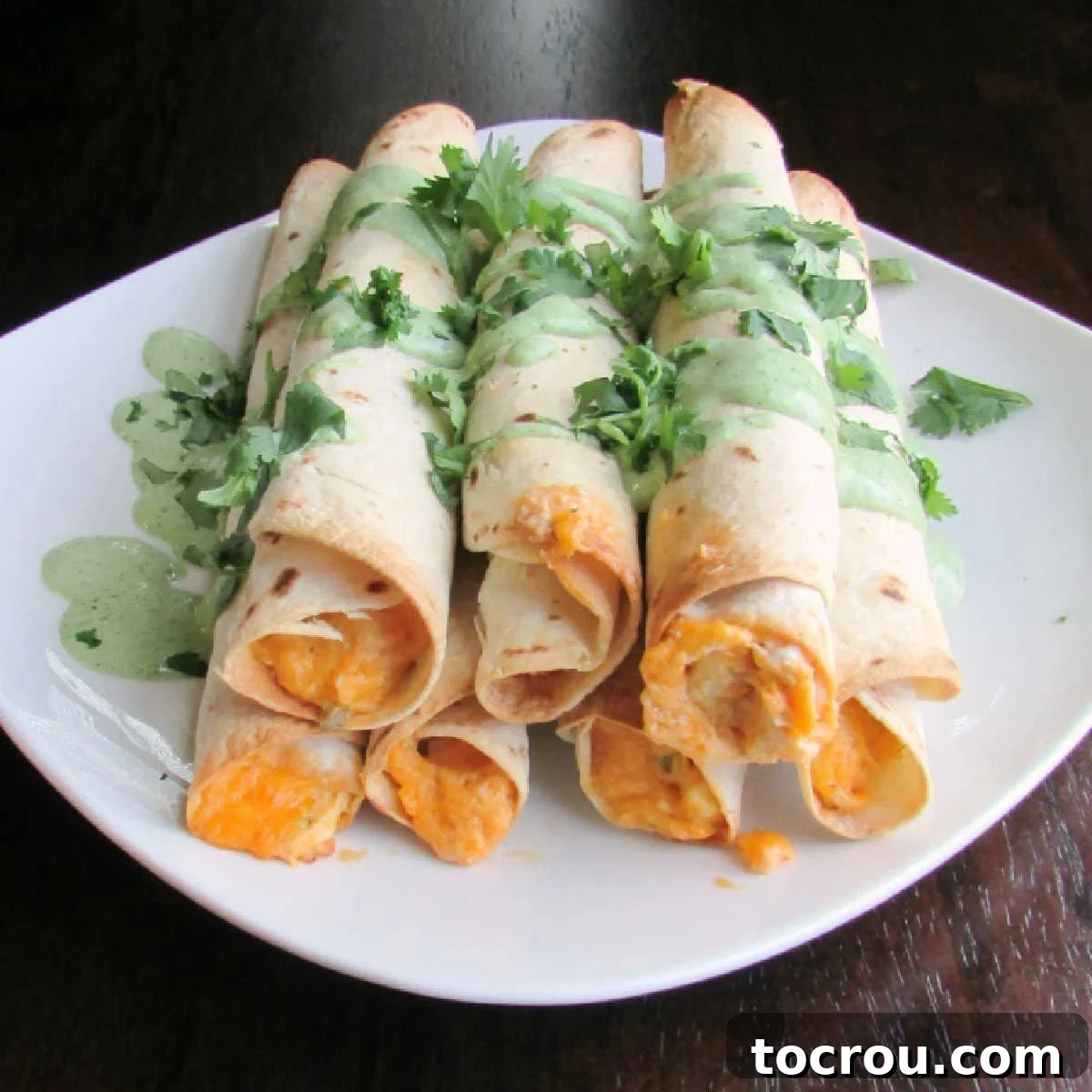 Stack of crispy baked buffalo chicken taquitos with fresh cilantro cream sauce and chopped cilantro garnish on top.
