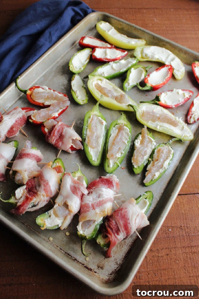 Jalapeno peppers in various stages of being stuffed, some with cream cheese, some with cream cheese and shrimp and some already wrapped in bacon with a toothpick holding it together on sheet pan.
