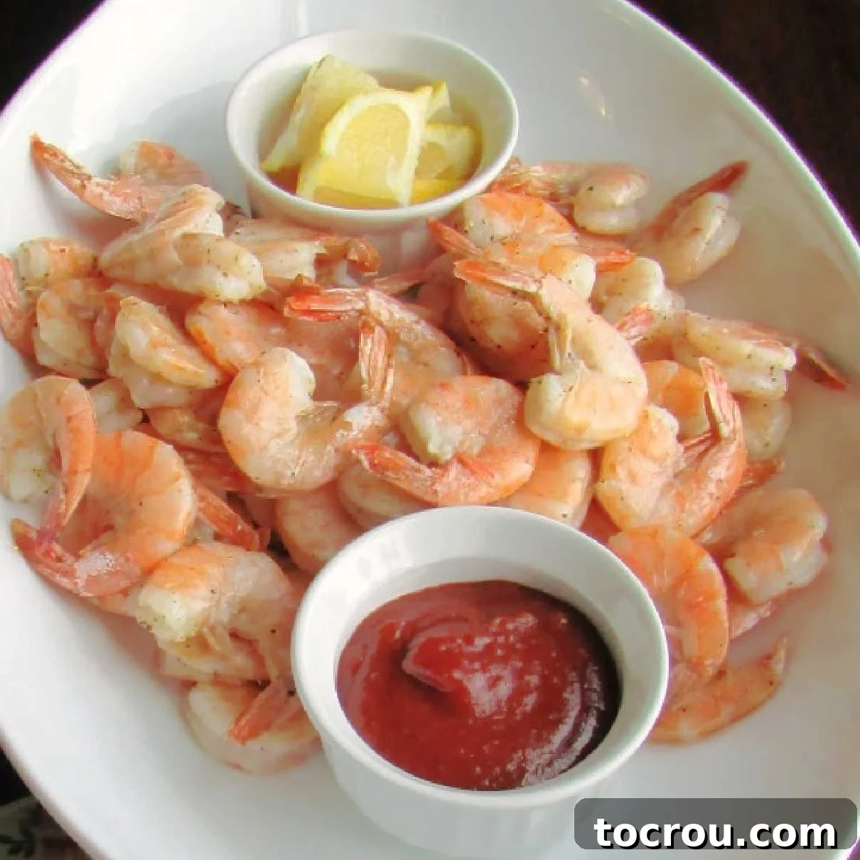A beautifully arranged platter of golden lemon roasted shrimp, accompanied by a small bowl of classic cocktail sauce and fresh lemon wedges, ready to be served.