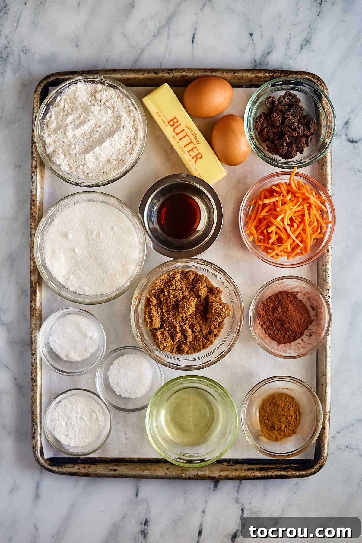 Ingredients including raisins, shredded carrots, flour, sugar, butter, eggs, vanilla, cocoa powder, brown sugar and more ready to be made into carrot cake.