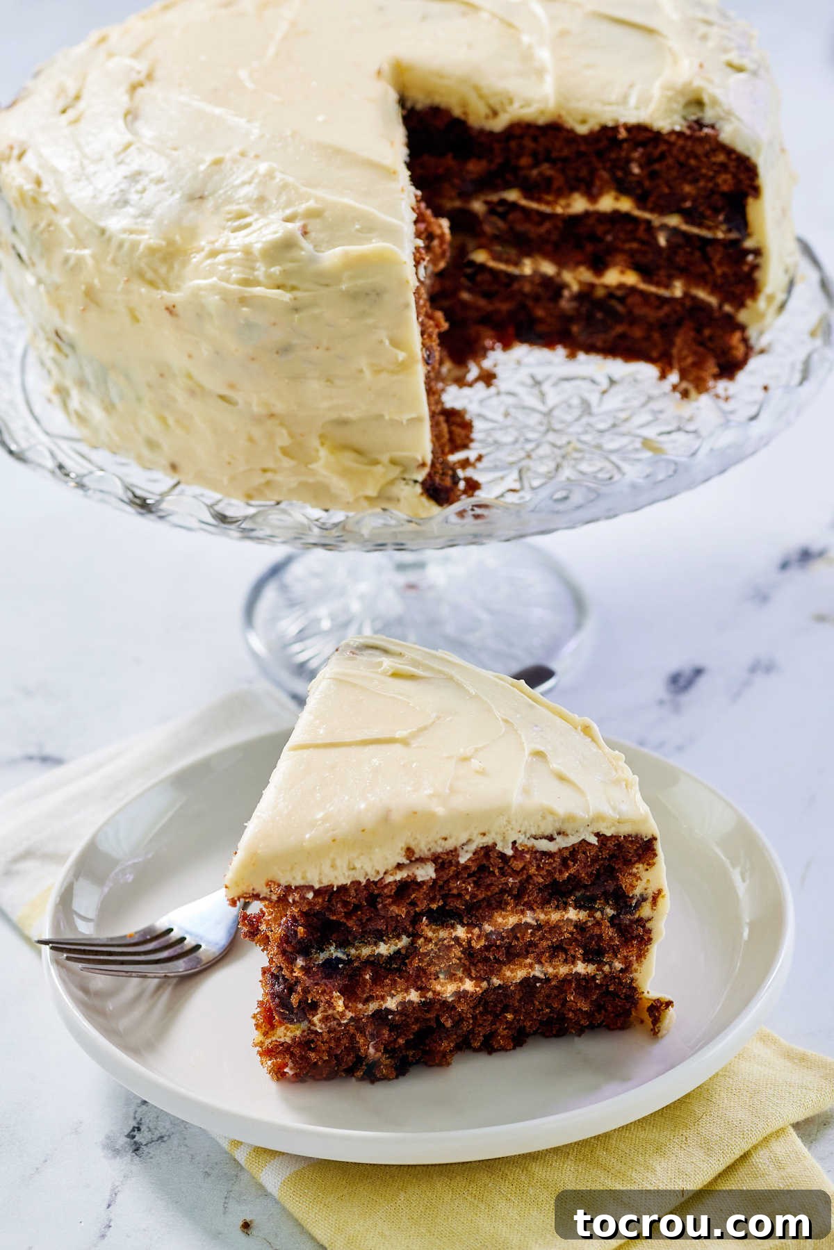 Large slice of layered carrot cake with cream cheese frosting on a plate with remaining cake on cake stand in the background.