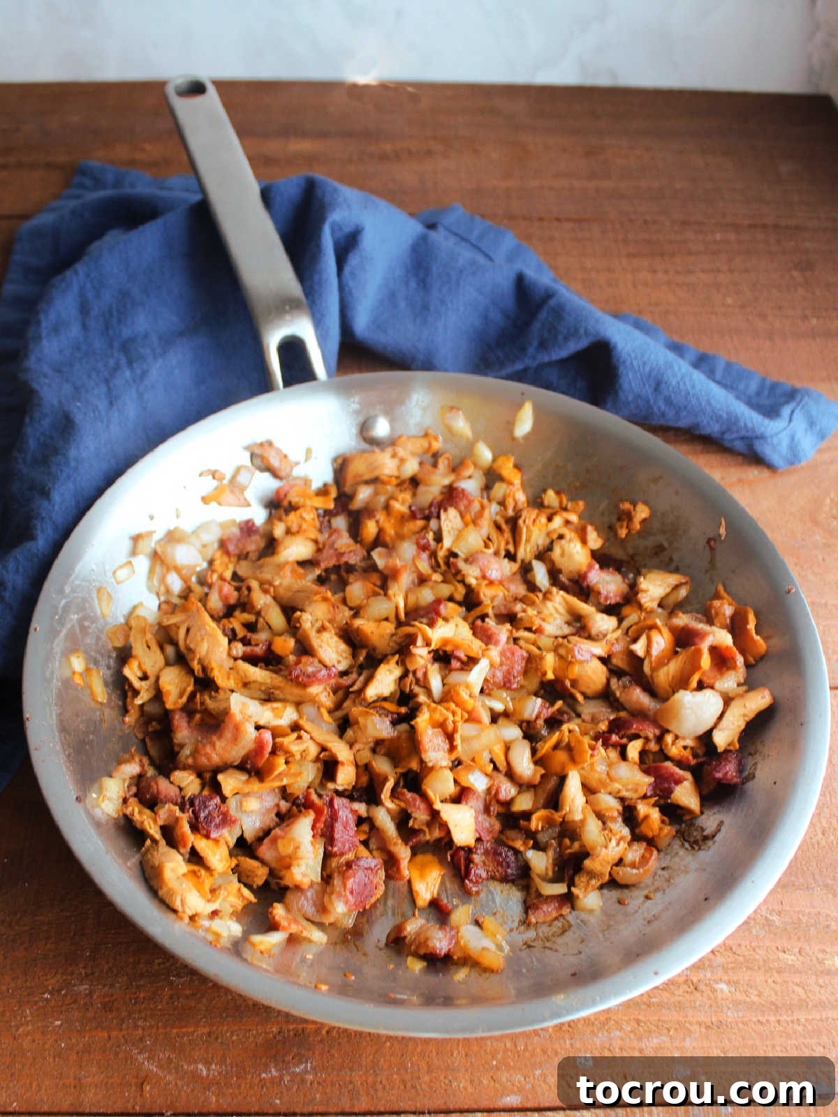 Stainless steel skillet featuring sautéed chanterelle mushrooms, tender onions, and crispy bacon bits, creating an aromatic base for the creamy sauce.