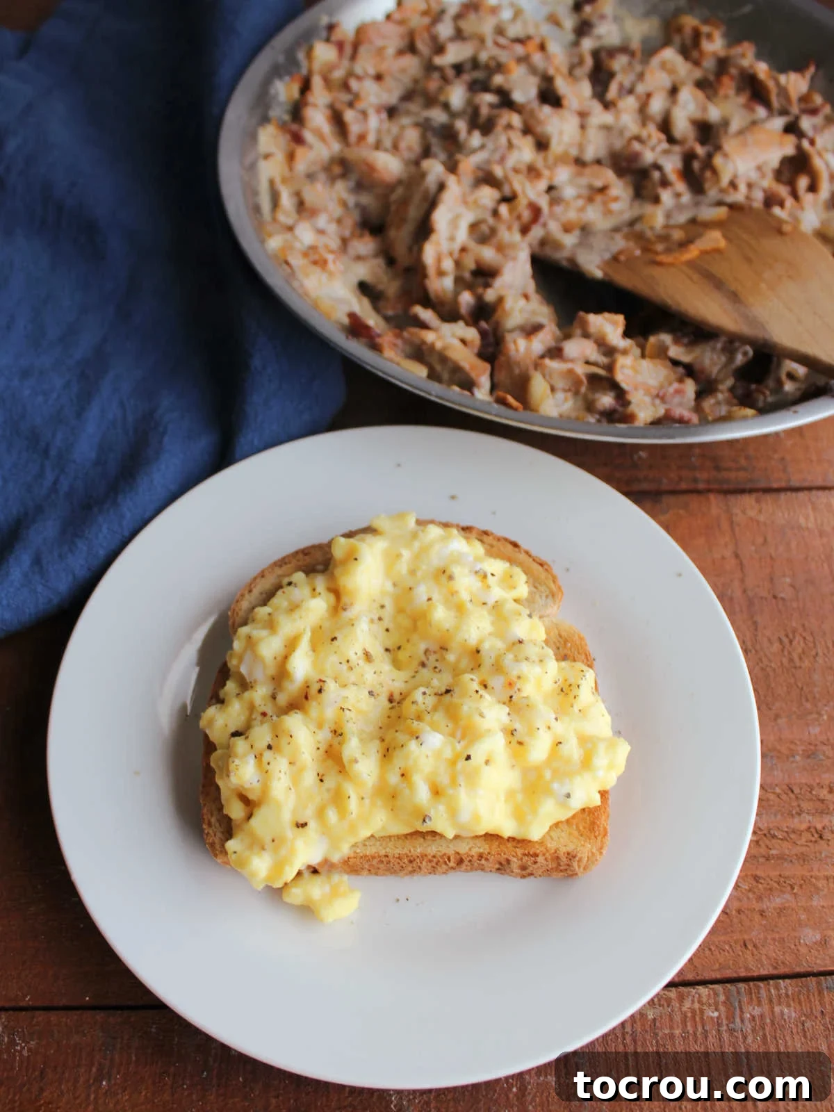 Plate with a piece of golden toast topped with soft scrambled eggs and fresh ground black pepper, with a skillet of creamy chanterelle and bacon mixture blurred in the background, awaiting its topping.