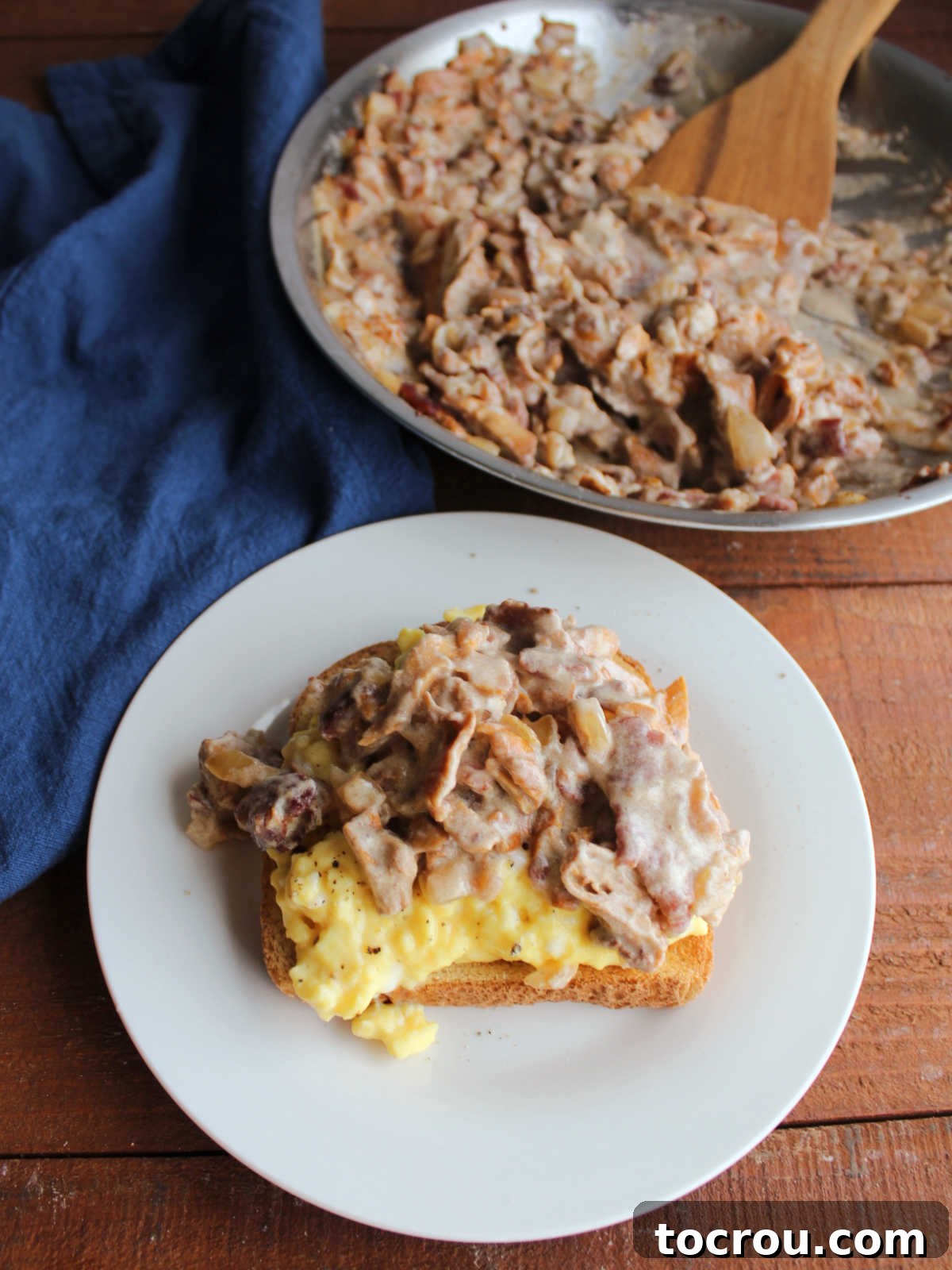 A plate featuring creamy Russian chanterelle and bacon mixture served generously over a piece of toast and soft scrambled eggs, with the remaining mushroom mixture visible in a skillet in the background.