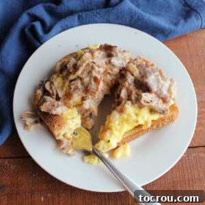 Fork getting a bite of toast topped with soft scrambled eggs and creamy russian chanterelle mushrooms with bacon and onions.