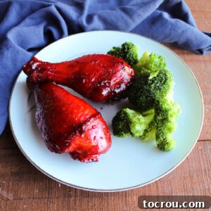 Dinner plate with two glossy red char siu chicken drumsticks and steamed broccoli, ready to eat and garnished with sesame seeds.