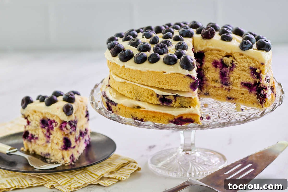 A beautifully presented slice of lemon blueberry layer cake on a white plate, positioned next to the majestic full cake on a pedestal. The cake displays multiple layers, vibrant blueberries, and luscious cream cheese frosting, inviting immediate indulgence.