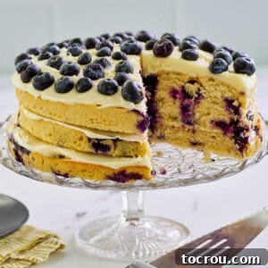 A grand, multi-layered lemon blueberry cake standing proudly on a cake stand, revealing its moist layers baked with fresh blueberries and thick, creamy cream cheese frosting in between. A slice has been removed, showcasing the beautiful interior texture and inviting everyone to taste.