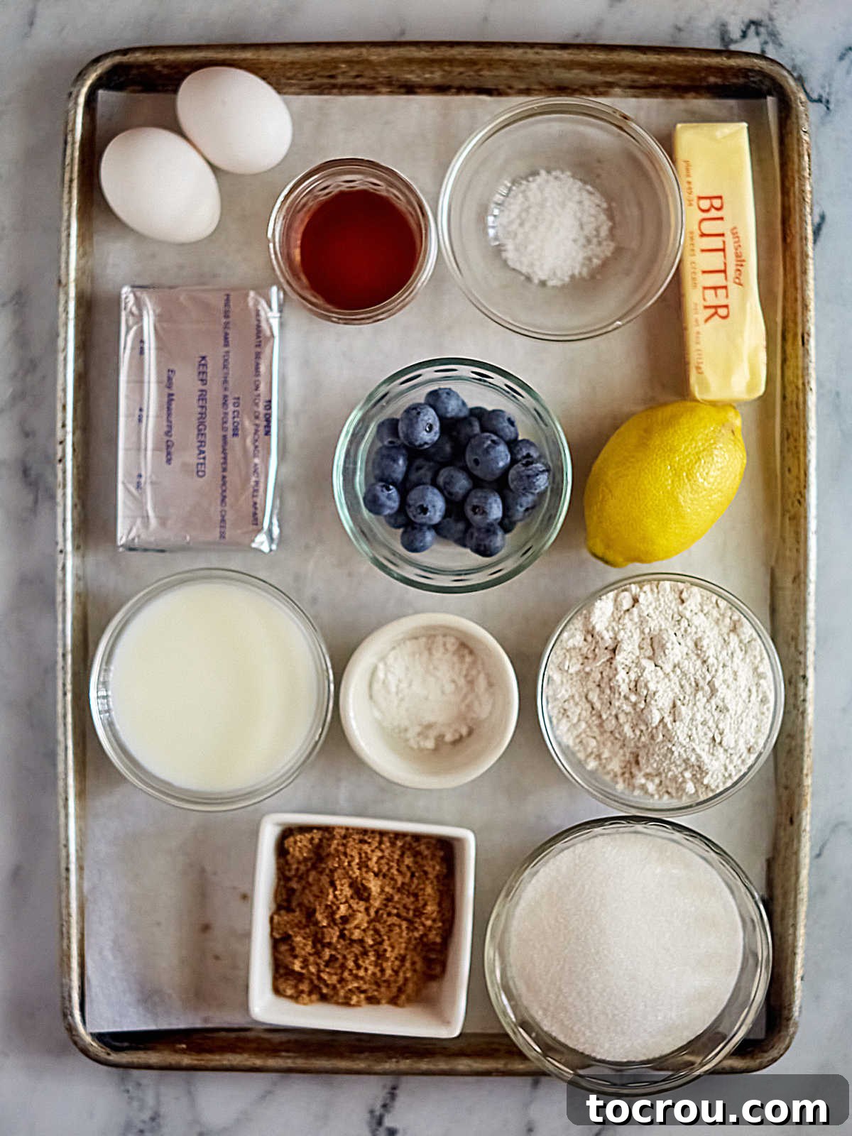 A vibrant display of fresh ingredients for baking a lemon blueberry layer cake, including ripe lemons, plump blueberries, all-purpose flour, granulated sugar, softened butter, cream cheese, and fresh eggs, meticulously arranged on a countertop, ready for preparation.