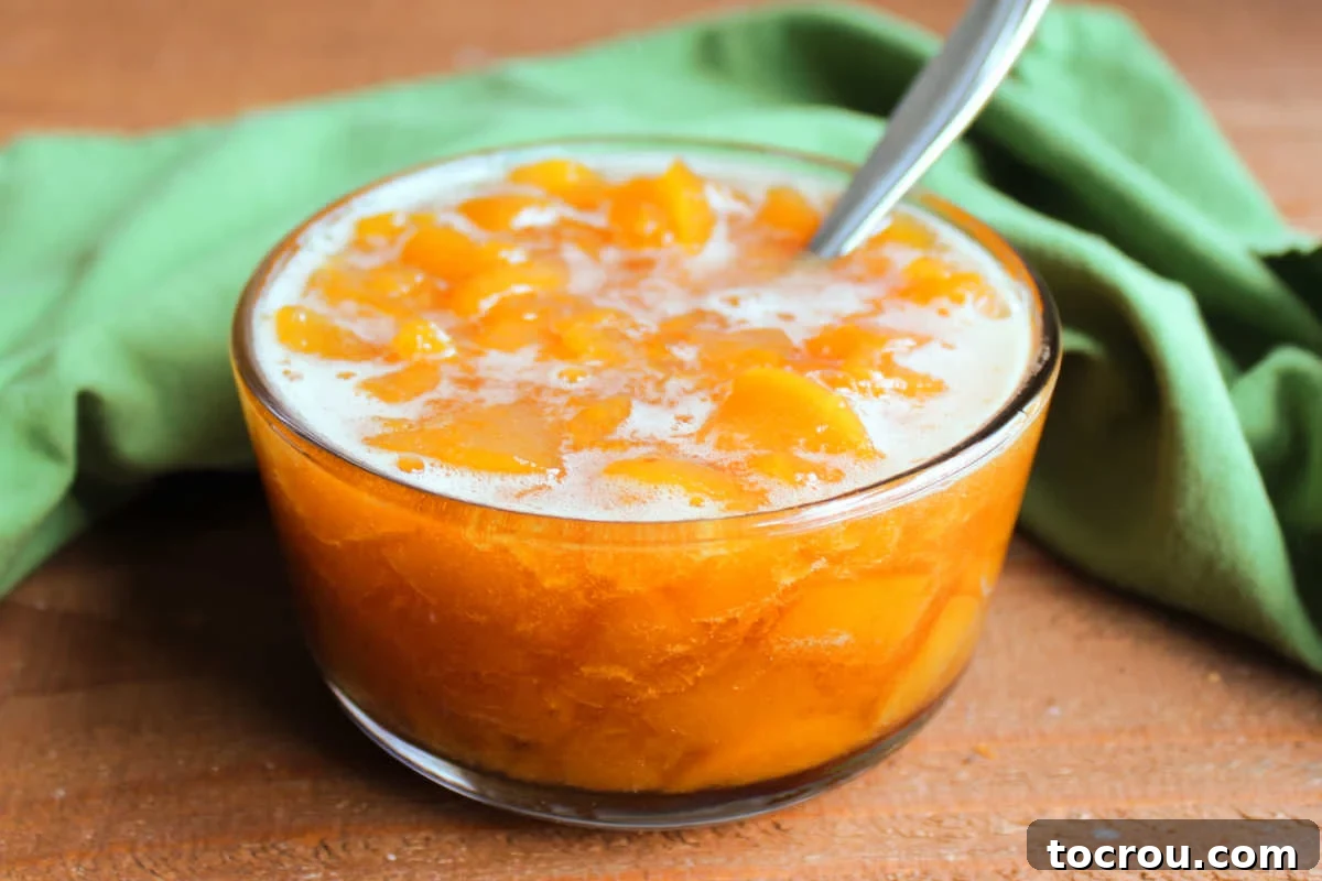 Homemade Peach Sauce A glistening glass bowl filled with homemade peach sauce, showcasing generous chunks of perfectly cooked peaches in a rich, thickened syrup.