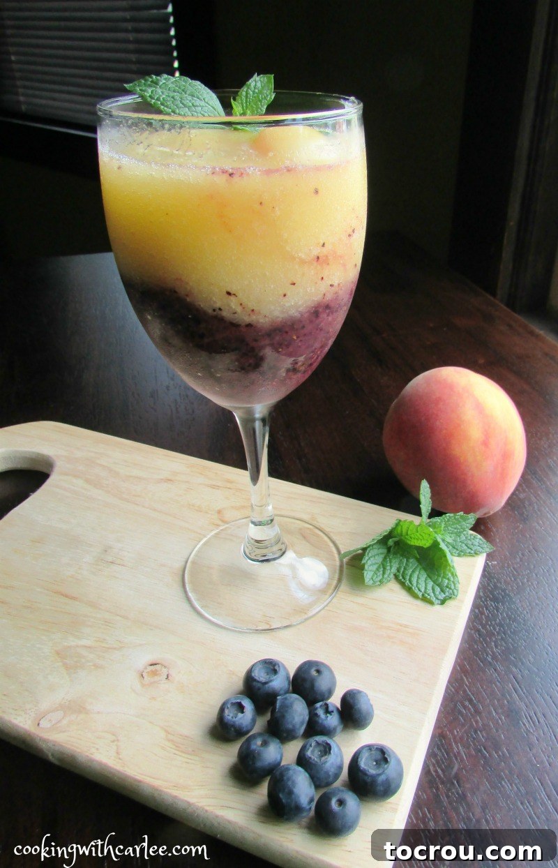 A single wine glass showcasing a perfect layer of vibrant blueberry wine slush at the bottom, topped with a smooth, creamy peach wine slush. Fresh mint leaves add an elegant garnish.