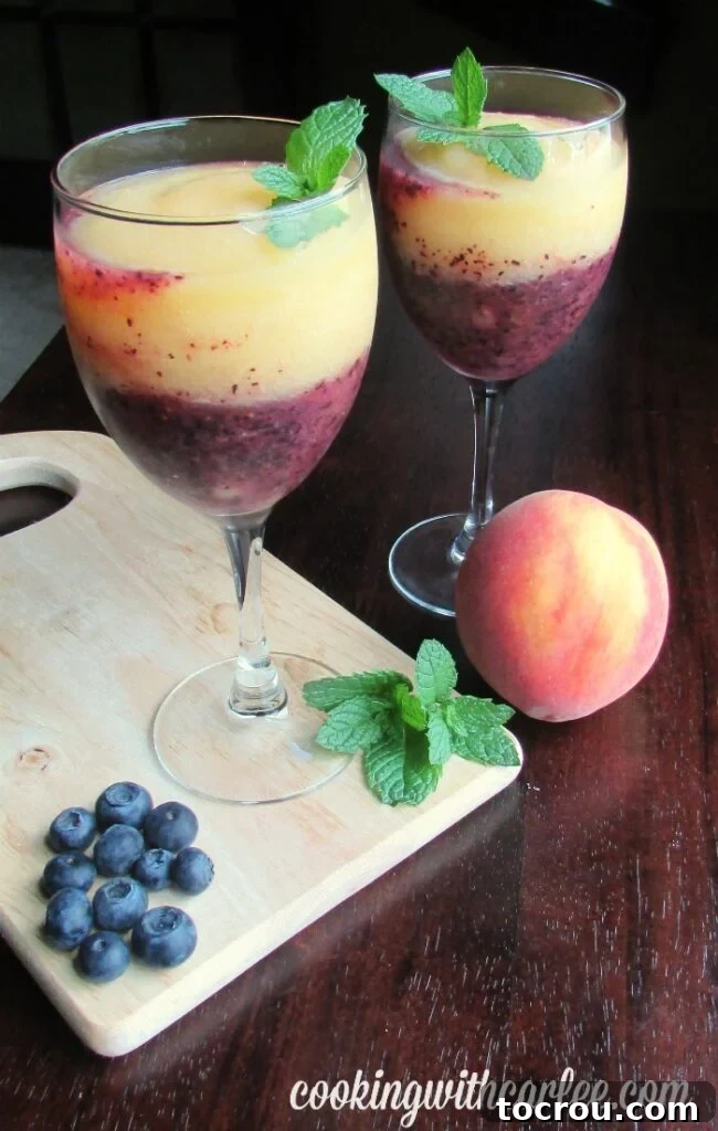Two elegant wine glasses, each filled with distinct layers of deep blue blueberry wine slush and light orange peach wine slush, adorned with fresh green mint sprigs.