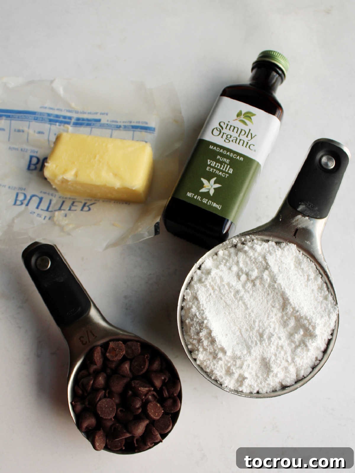 Chocolate Fudge Cream Delight 6 Ingredients for chocolate icing, including shiny chocolate chips, softened butter, powdered sugar, and a bottle of vanilla extract, artfully arranged on a clean surface.