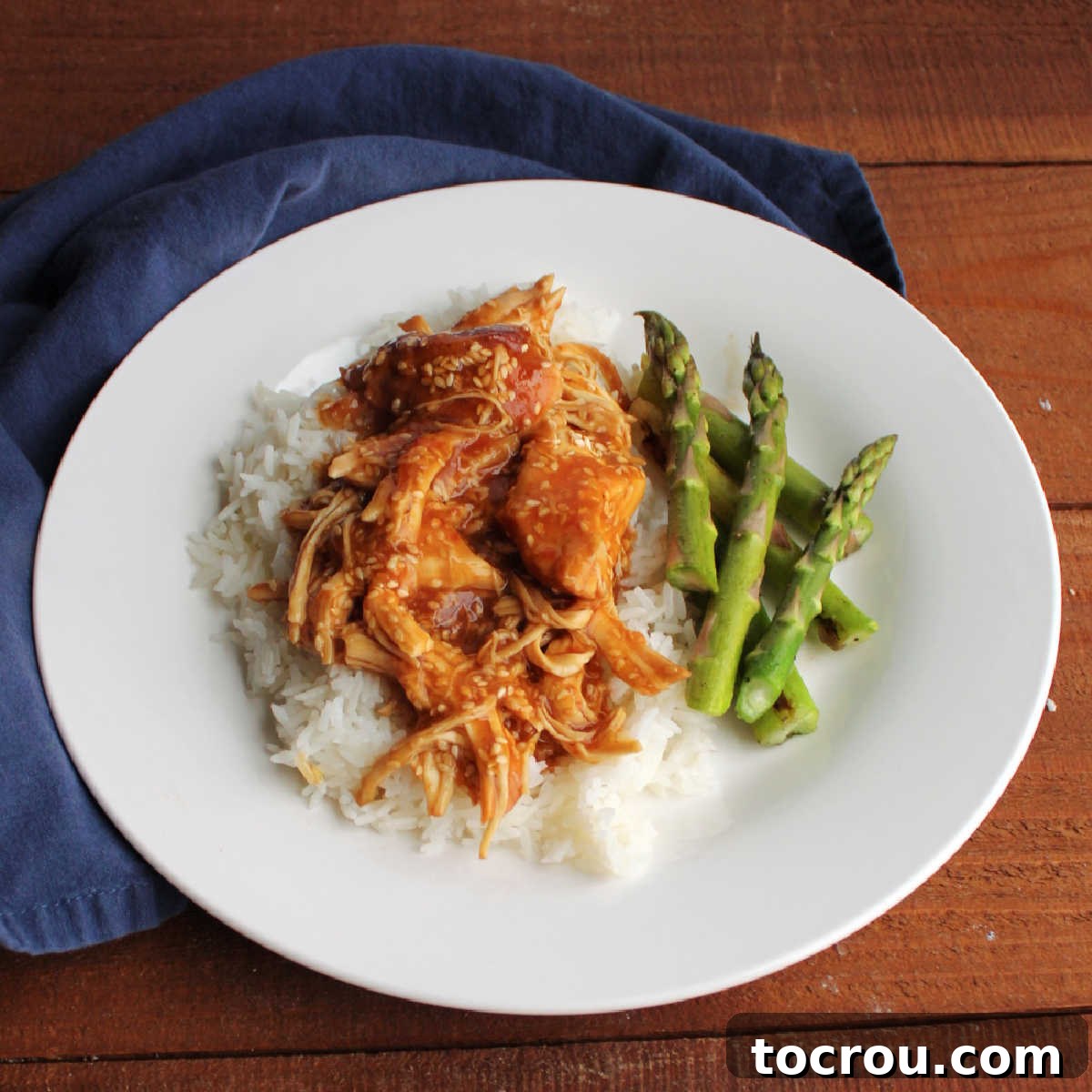 A dinner plate featuring a hearty portion of slow cooker sesame chicken over rice, accompanied by perfectly roasted asparagus spears.