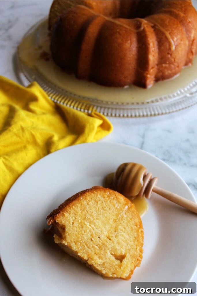 A slice of glistening honey lemon bundt cake served on a plate, with a rustic wooden honey drizzler nearby, ready to add more sweetness.
