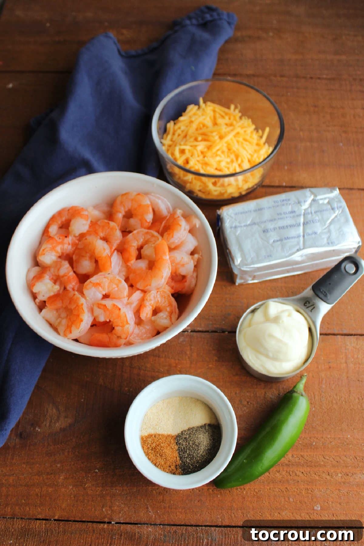 Array of fresh ingredients laid out for Cajun shrimp dip, including large shrimp, cream cheese, mayonnaise, a vibrant jalapeño, various spices, and shredded cheddar cheese.