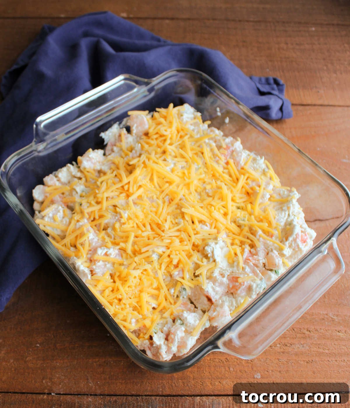 An 8-inch square baking dish filled with unbaked Cajun shrimp dip, generously topped with shredded cheddar cheese, ready for the oven.