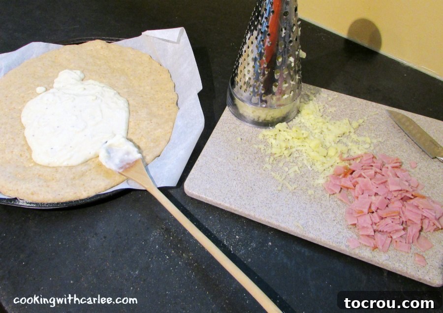 A smooth, round pizza crust generously spread with creamy béchamel sauce, resting beside a wooden cutting board piled with diced ham and grated cheese, all ready for the next step in pizza assembly.