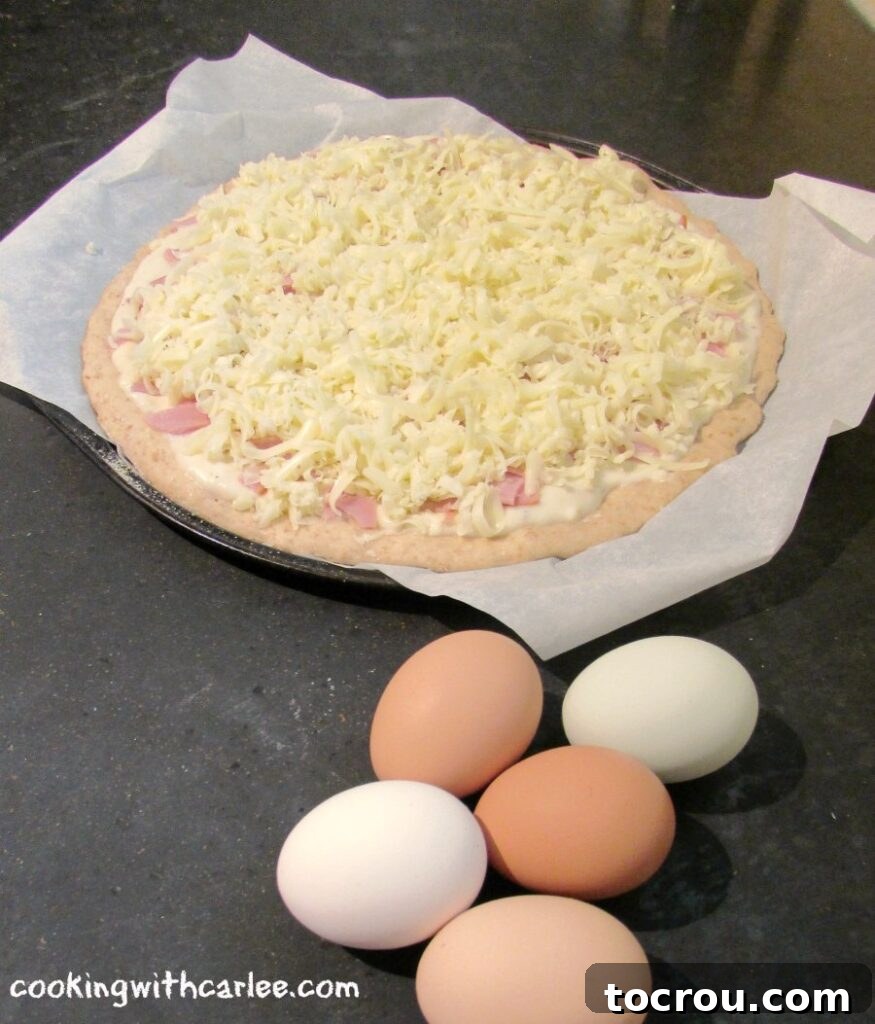Three rich brown eggs placed alongside an unbaked pizza, which is already topped with a creamy white sauce, diced ham, and a generous layer of mixed cheeses, patiently waiting to be baked.