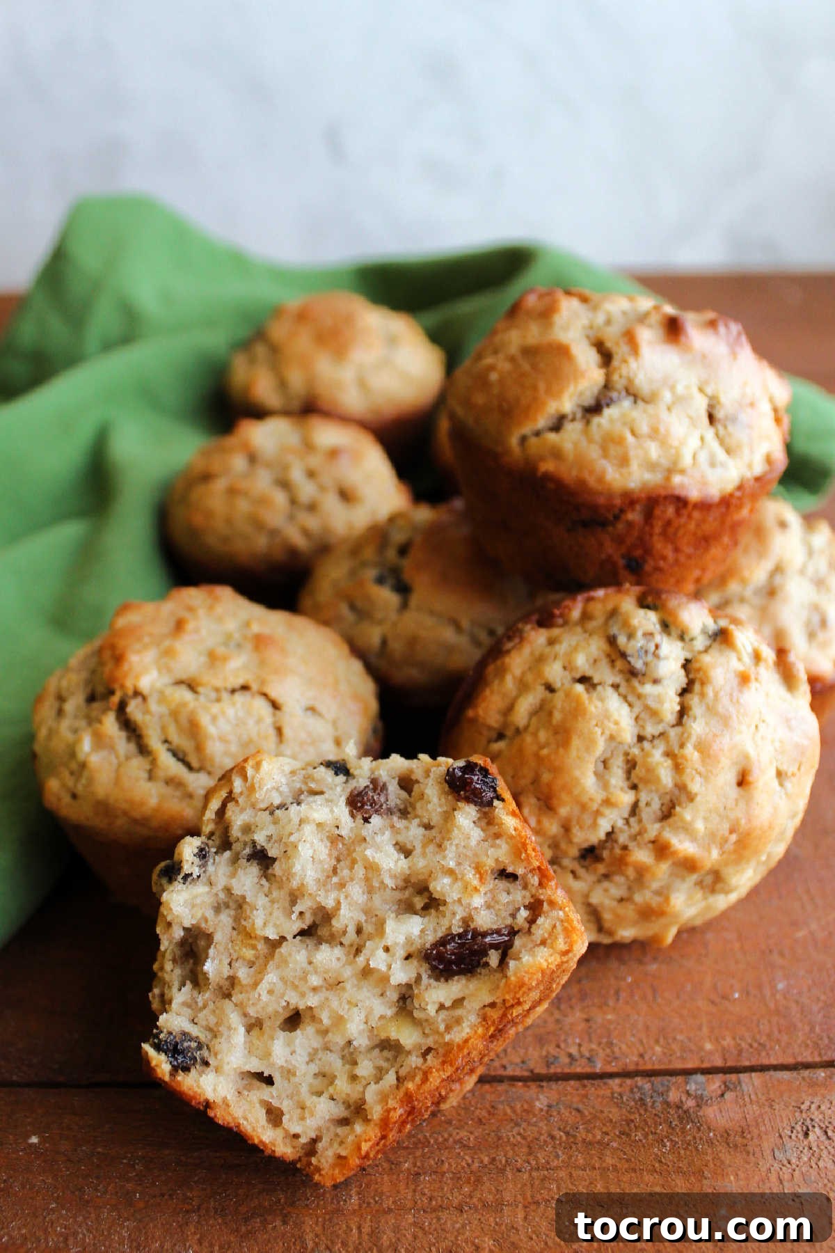 A close-up of a halved oatmeal raisin sourdough muffin, showing its moist, tender interior with visible oats and raisins, and more whole muffins in the background.