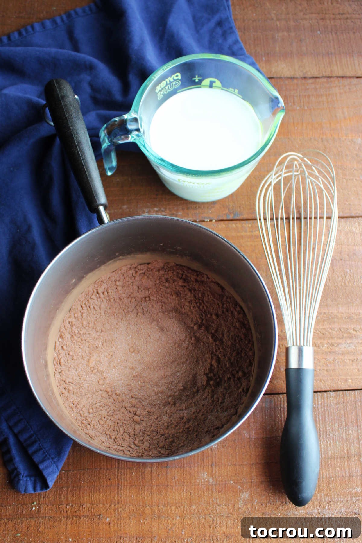 Velvet Mocha Cloud Frosting 5 Sugar, flour, cocoa powder and instant coffee stirred together in saucepan with whisk and measuring cup of milk nearby.