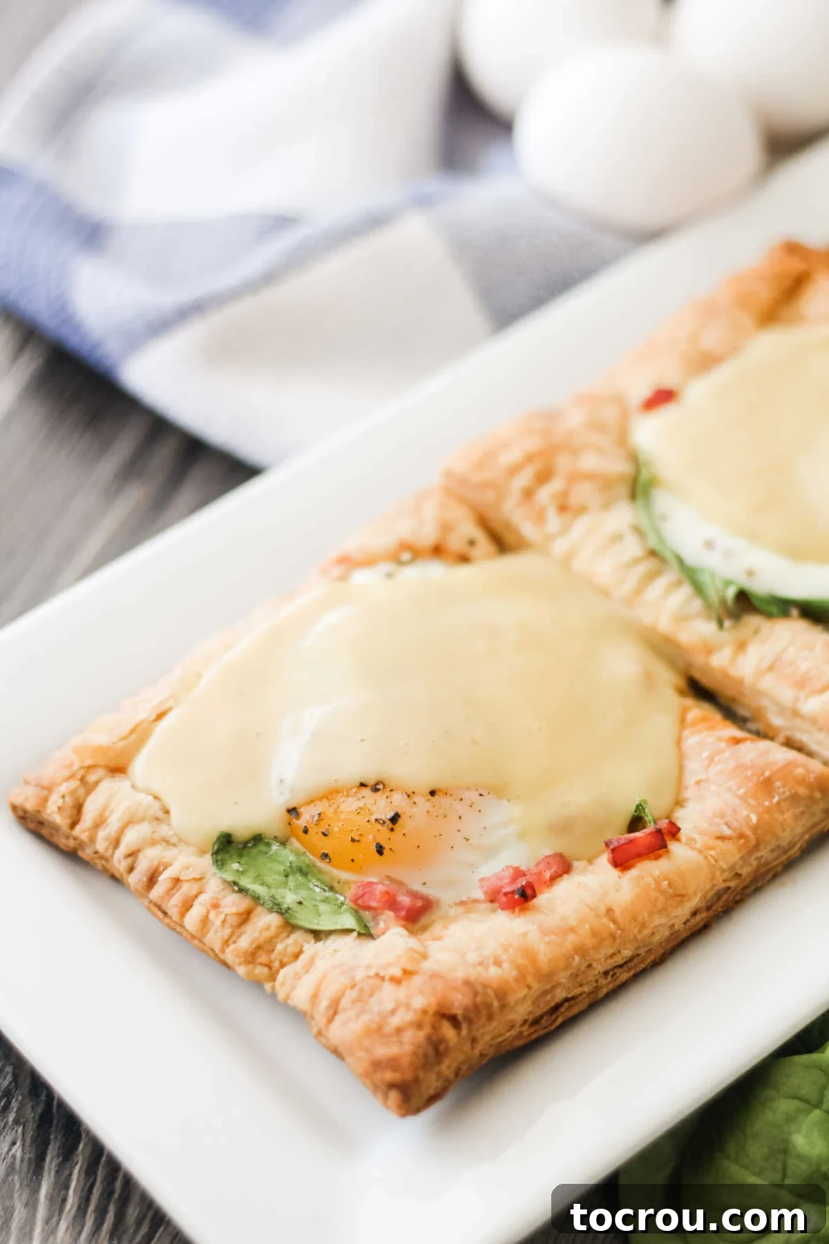 Close-up of a puff pastry tart, generously topped with cooked ham, vibrant spinach, a perfectly baked egg, and a rich drizzle of hollandaise sauce.
