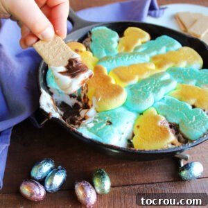 Child's hand holding piece of graham cracker dipped into smore dip with gooey melted Peeps on top of melted chocolate mixture.