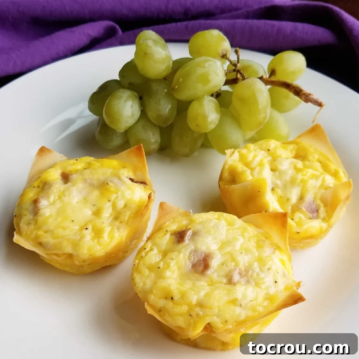 Delicious mini quiches with grapes for breakfast Three golden brown mini ham and cheese quiches in crispy wonton wrappers, served on a white plate alongside fresh green grapes.