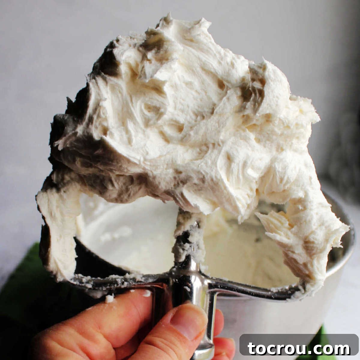 Hand holding stainless steel mixer paddle filled with fluffy white crusting buttercream, showcasing its smooth texture.