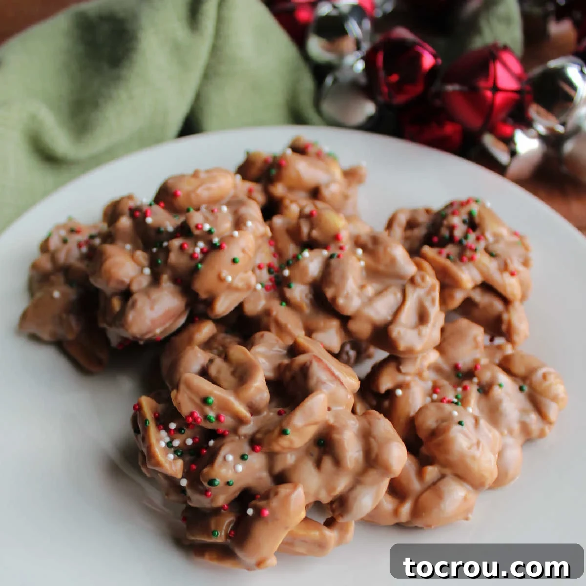 A deliciously chocolaty treat, these peanut clusters are made in a slow cooker. They couldn't be simpler to make and while they cook you can read the Christmas poem MiMi wrote! Chocolate peanut clusters topped with Christmas sprinkles on plate, ready to eat.