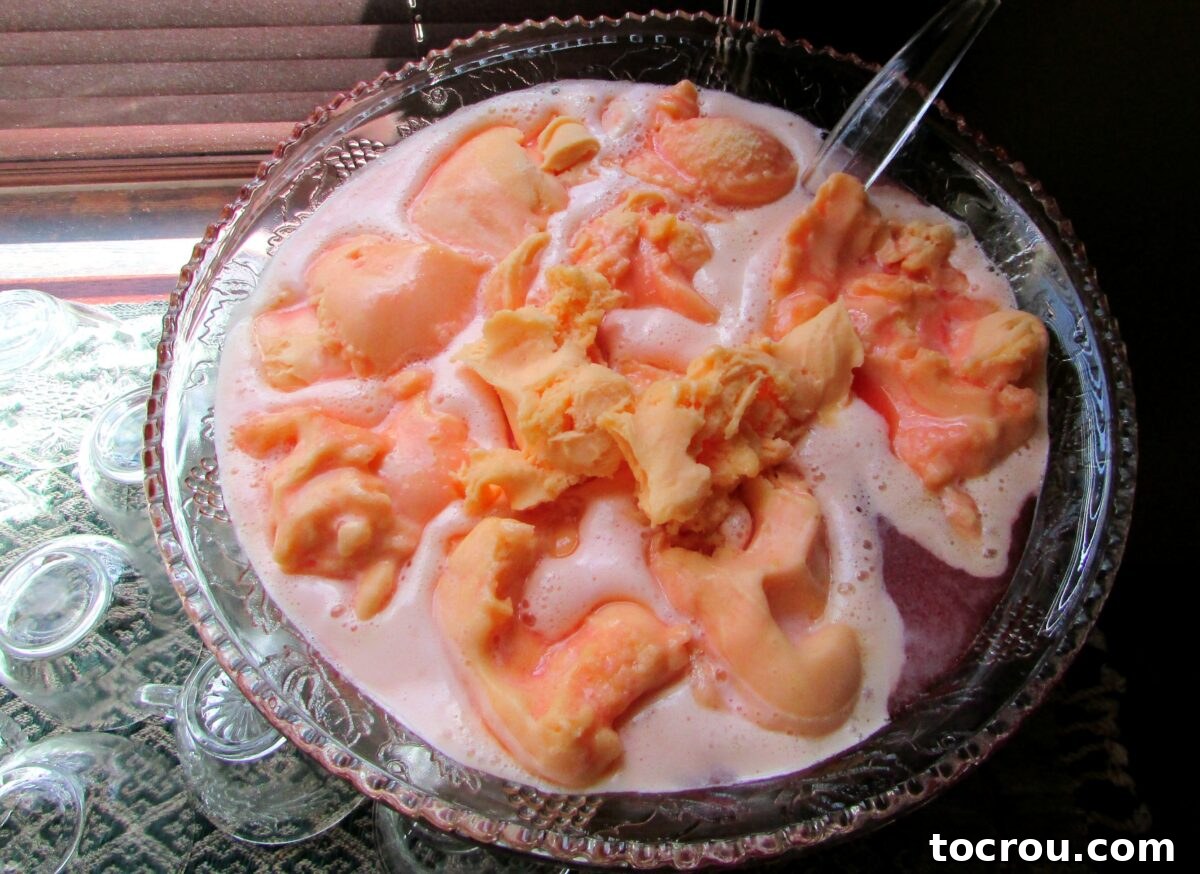 large glass punch bowl filled with pink punch topped with scoops of orange sherbet.