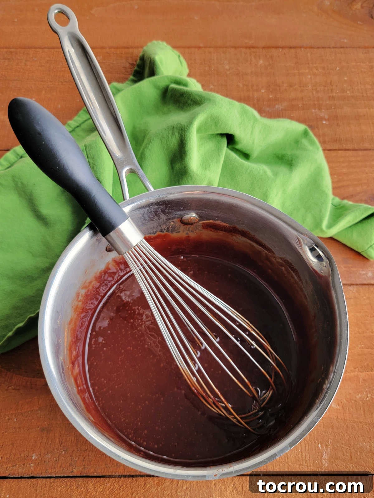 Freshly cooked fudge icing glistening in a saucepan with a whisk resting in it, ready to be poured over a dessert.