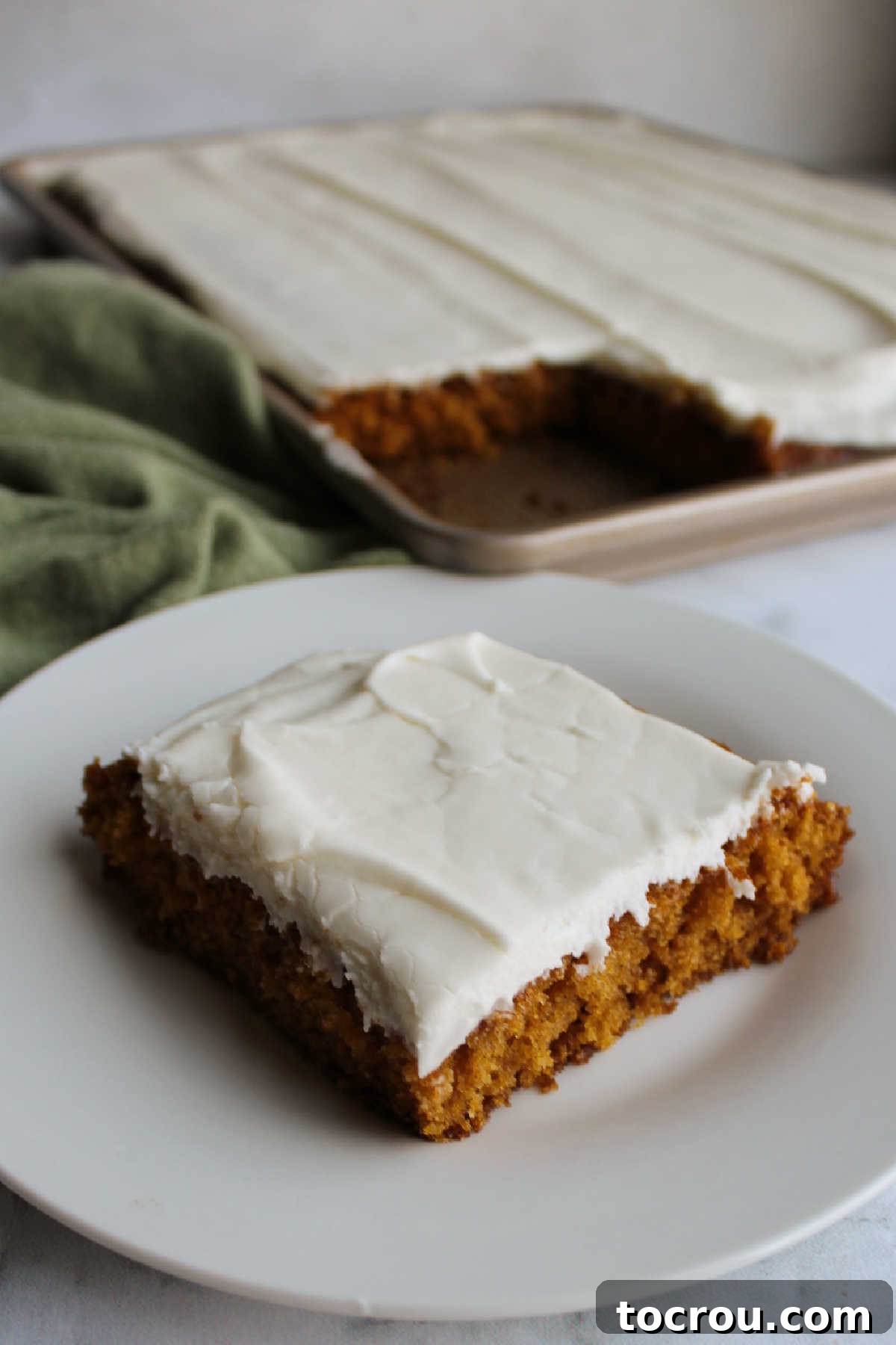 A single, perfectly frosted pumpkin bar sits on a white plate in the foreground, with the remaining pan of delicious bars slightly blurred in the background.
