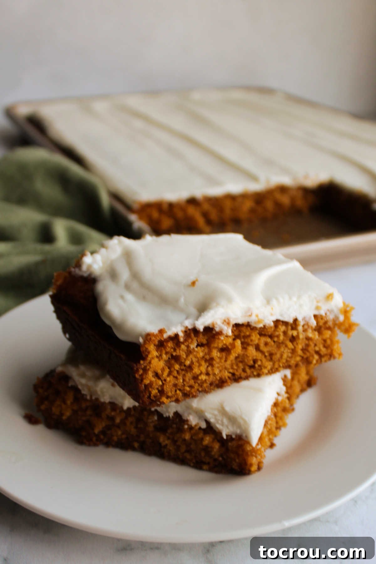 Several moist pumpkin squares with creamy frosting arranged on a white plate, positioned in front of the full sheet pan of bars.