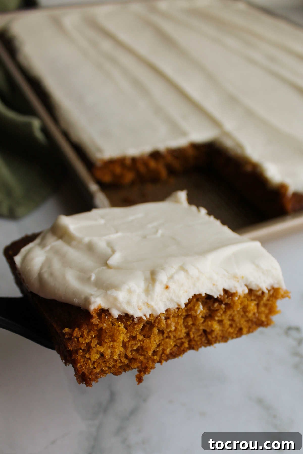 A freshly cut pumpkin square, generously topped with cream cheese frosting, being carefully lifted out of a large sheet pan with a spatula.