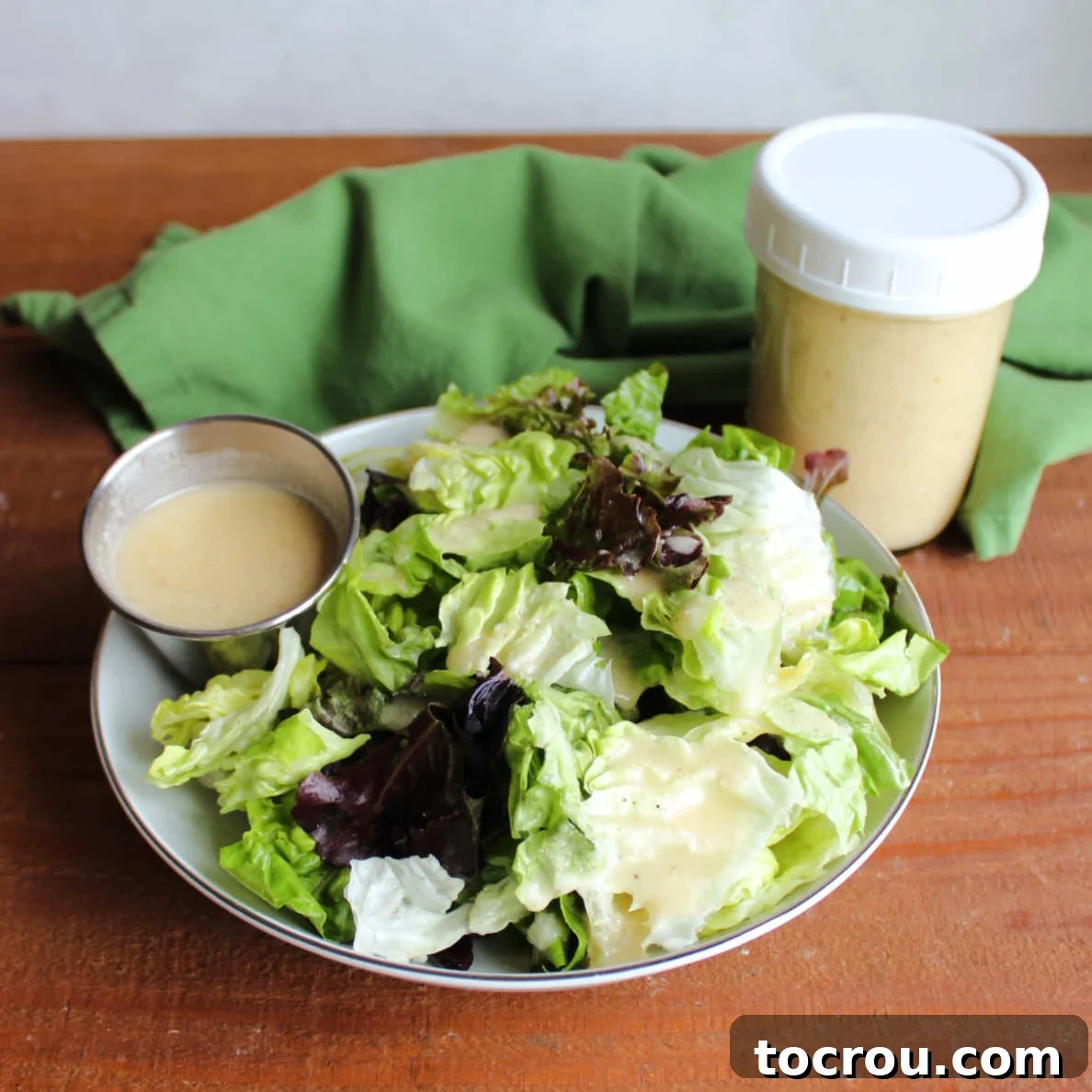 Bowl of lettuce with a ramekin of homemade salad dressing that looks creamy from the addition of Parmesan cheese next to a jar with more salad dressing inside.