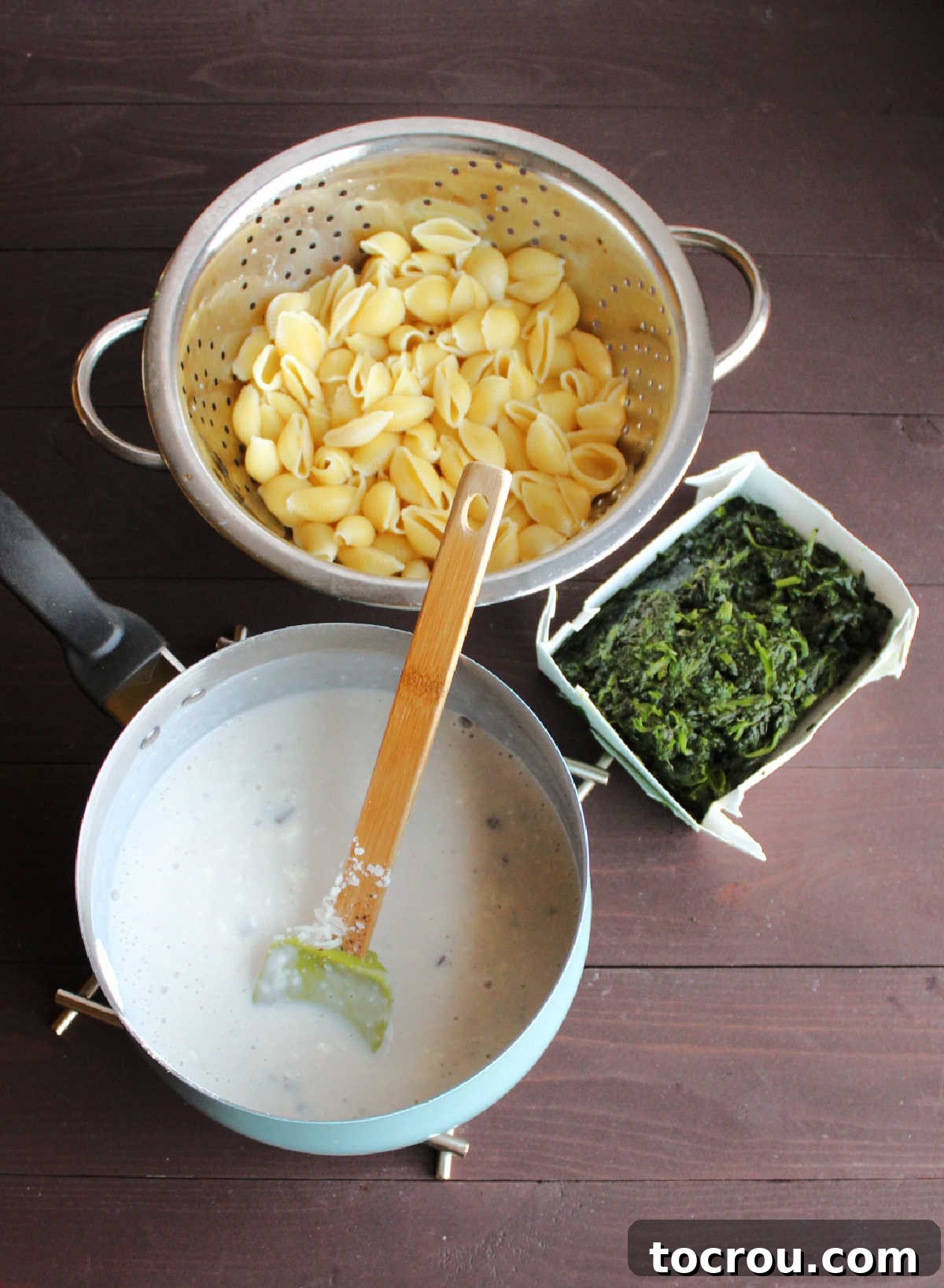 Velvety Spinach Macaroni Bake 3 Saucepan with creamy white cheese sauce, a colander full of cooked shell pasta, and a box of defrosted chopped spinach, ready for mixing.