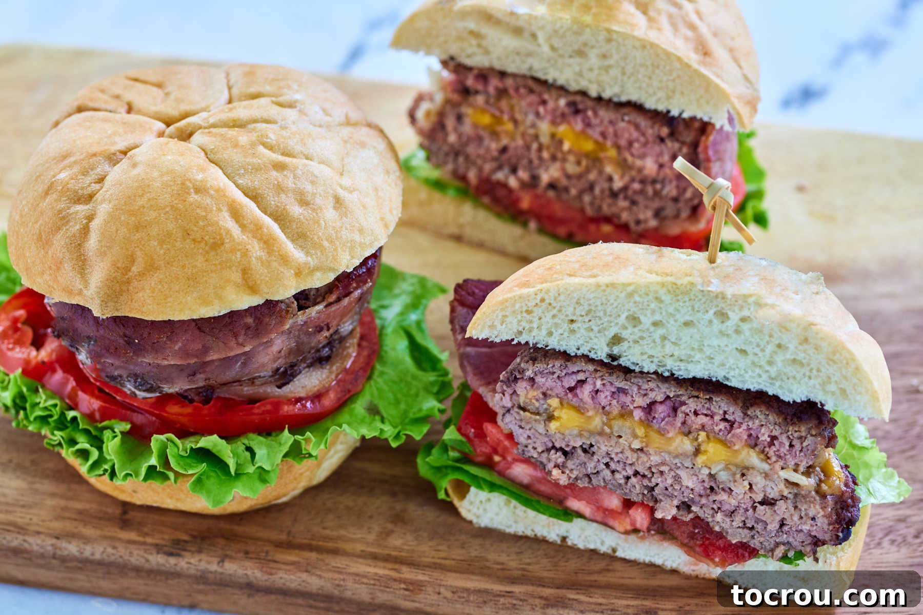 Maw-Maws Unexpected Burger Bliss 2 Whole hamburger surprise with bacon wrapped around it and tomato, lettuce and bun next to a burger that has been cut in half showing the filling of cheese, onions, and BBQ sauce. A culinary masterpiece ready to elevate any cookout.