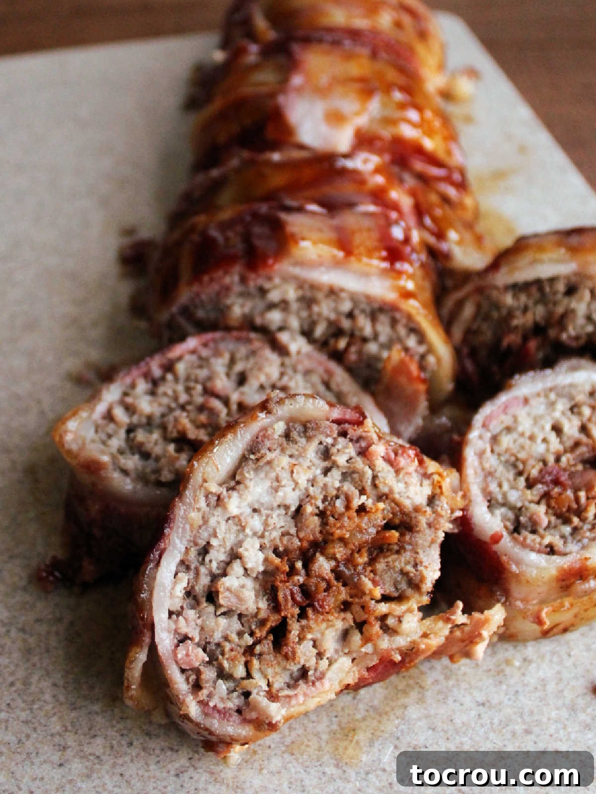 Cutting board with slices of bacon explosion showing crumbled bacon center, smoked ground pork and sausage layer and barbecued bacon weave exterior. 