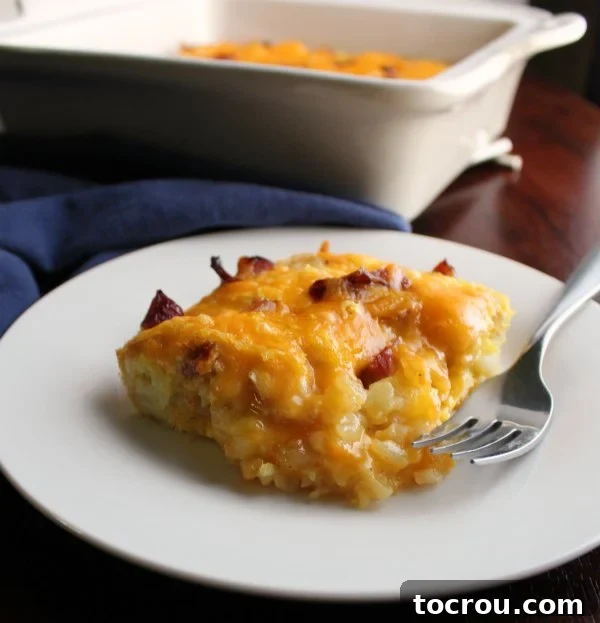 Tater Tot Casserole Ready to Eat A close-up shot of a piece of ham and tater tot breakfast casserole on a white plate, with a fork poised for a bite.