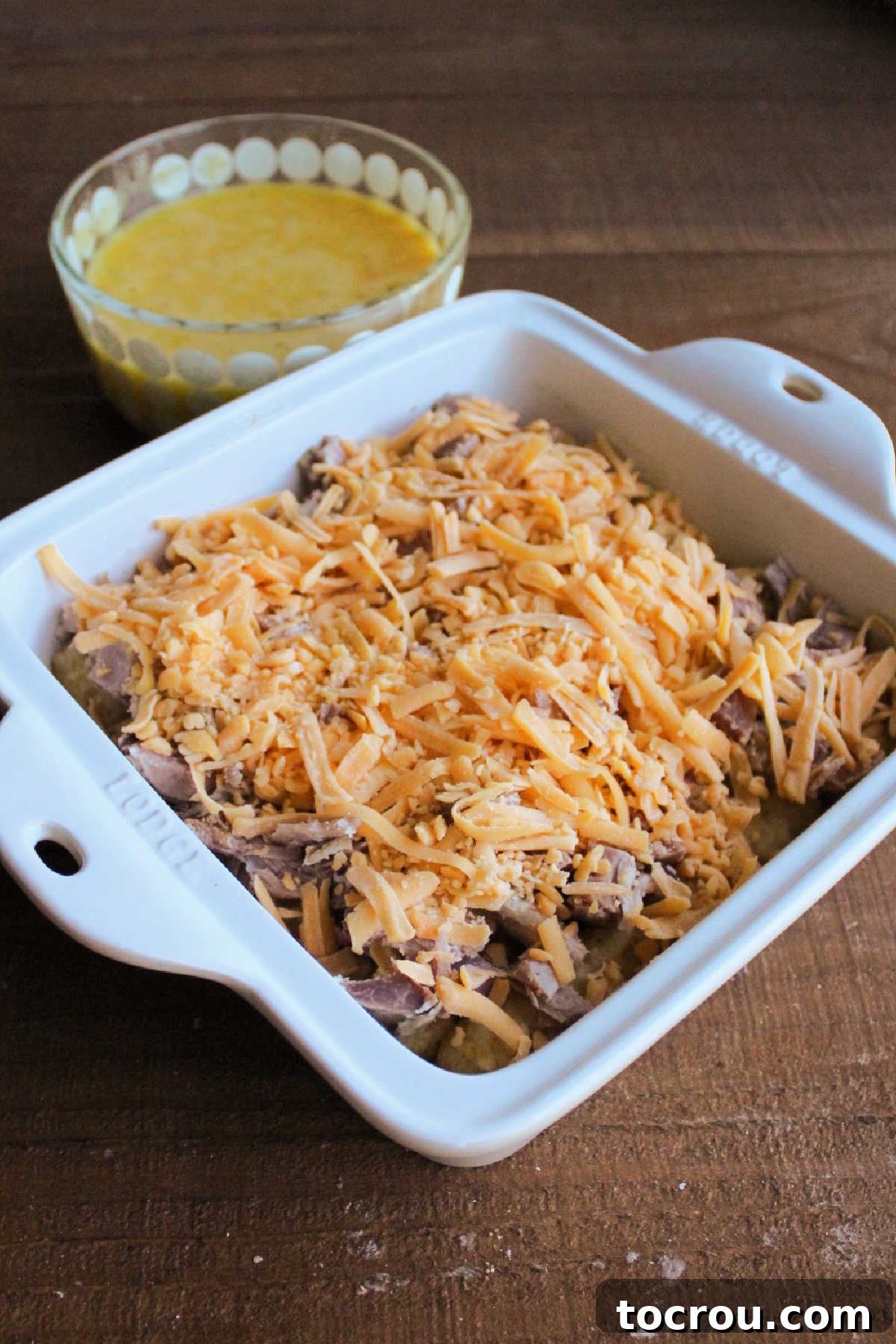 Layering Ingredients in Casserole A square baking dish filled with a base layer of tater tots, topped with generous chunks of ham and shredded cheese, with a bowl of beaten eggs in the background.