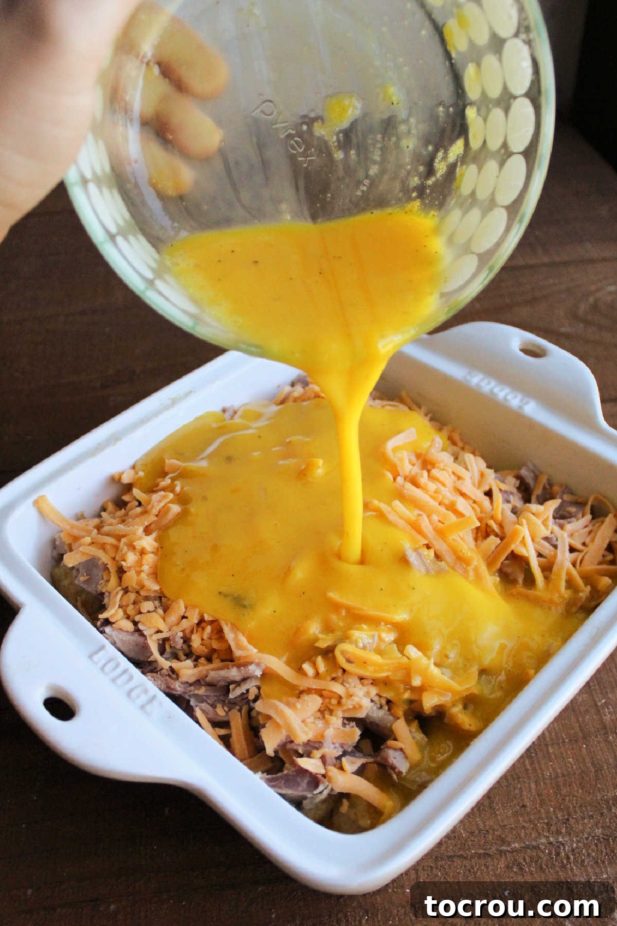 Pouring Eggs into Casserole The moment of pouring a bowl of perfectly seasoned beaten eggs over the arranged ham and cheese layers in a baking dish, preparing it for the oven.