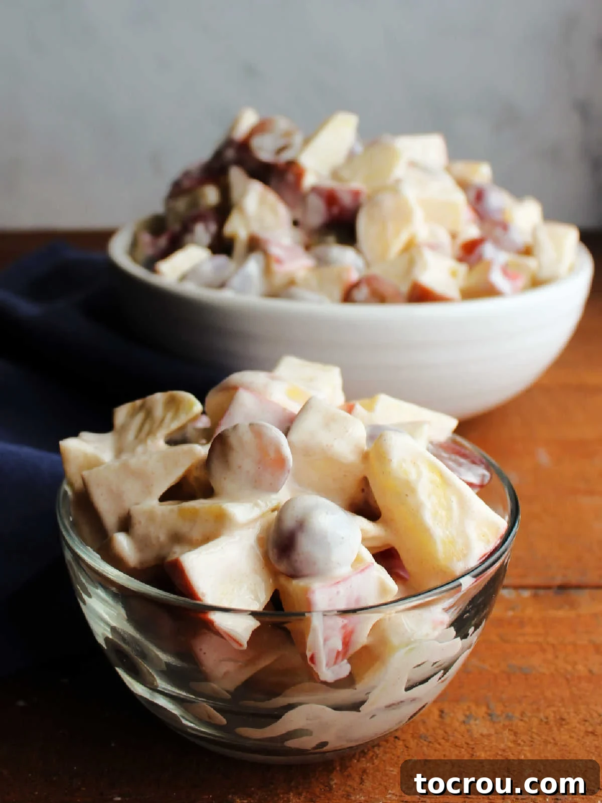 Individual and serving portions of apple and grape salad. Small glass bowl of grape and apple salad in front of large serving bowl with more fruit salad.