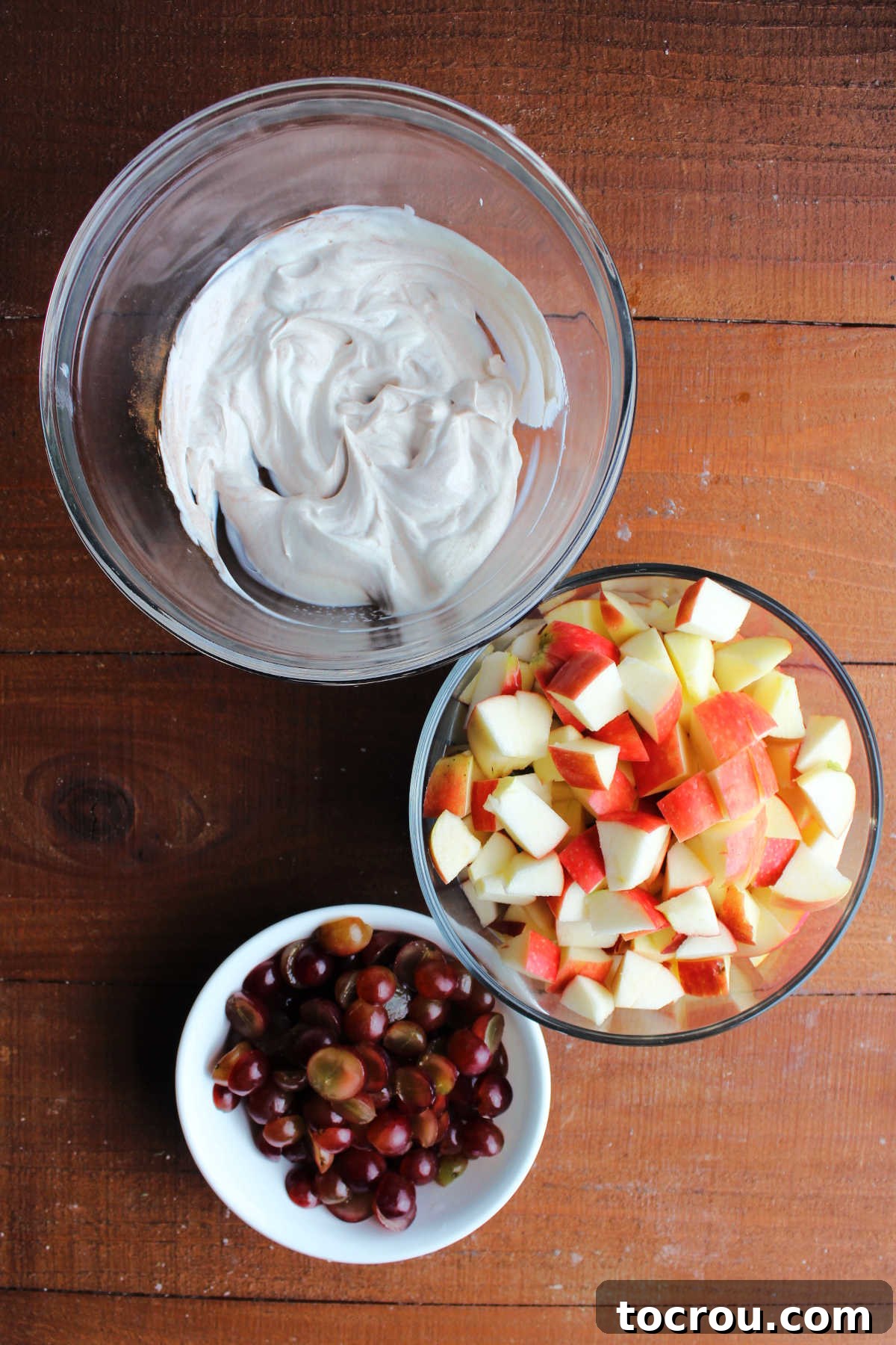 Preparation of apple and grape salad ingredients. Large mixing bowl with yogurt dressing next to a bowl of chopped apples and another bowl of halved grapes.
